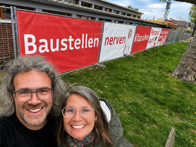 Arne und Adina vor dem Bauzaunbanner der Arbeitgebermarke am Kita-Neubau